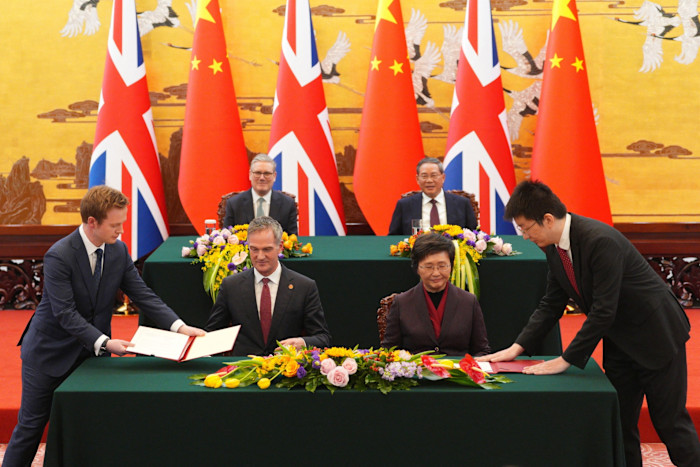 Keir Starmer and Li Qiang observe as Peter Kyle and Wang Xiaoping sign documents at a table decorated with flowers, with British and Chinese flags behind them.