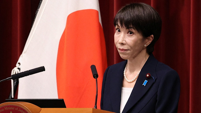 Sanae Takaichi stands at a podium, reacting during a press conference, with a Japanese flag in the background.