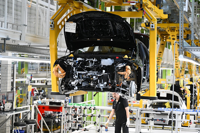 A worker assembles a Mercedes-Benz CLA sedan on an automated line, with the car’s hood open and machinery visible in the background.