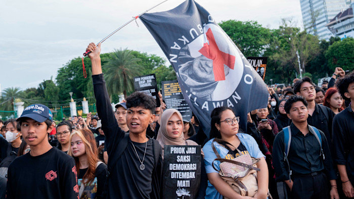 Protesters, many dressed in black, hold signs and a large flag during outside Indonesia’s presidential palace