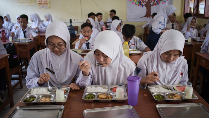 Students in uniform eat lunch from metal trays at their desks in a crowded junior high school classroom