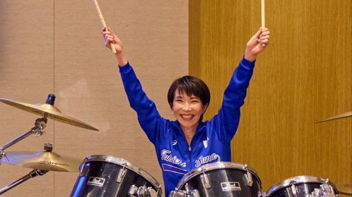 Sanae Takaichi smiles and raises drumsticks while seated at a drum kit, wearing a blue jacket with her name on it.