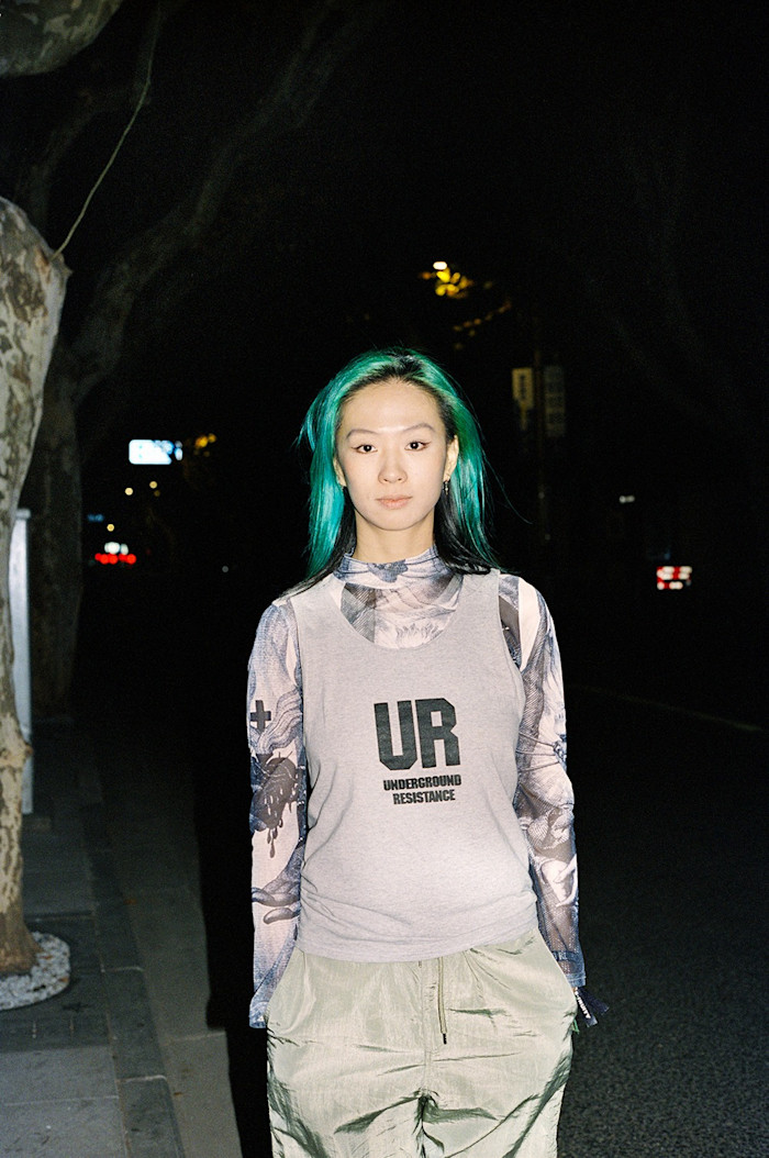A young woman with green hair wearing a "UR Underground Resistance" T-shirt smiles at the camera. She is up-and-coming DJ Liu Naxin, who is playing a set at Fruityshop Records