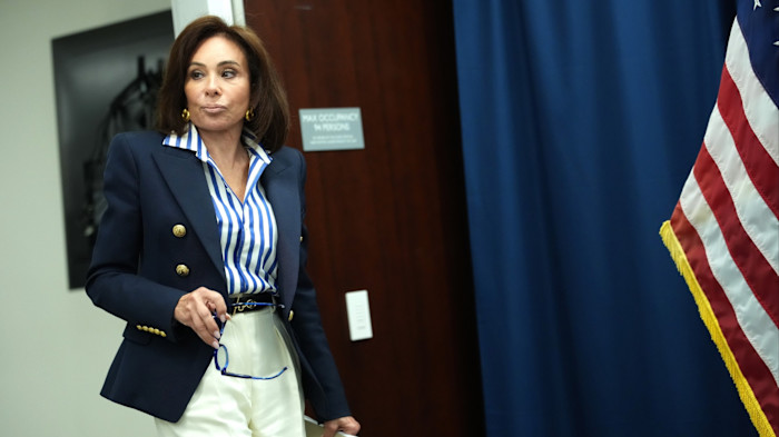 Jeanine Pirro enters a room, holding eyeglasses, ahead of a press conference with a US flag visible beside her.