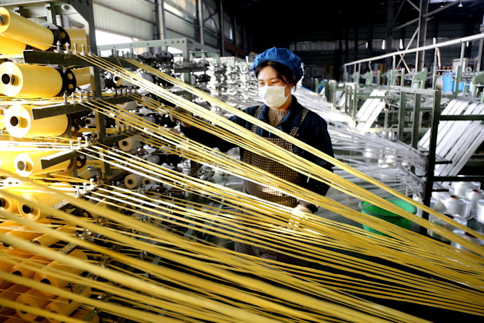 A worker wearing a mask and blue cap monitors yellow threads on weaving machines at a bag production workshop.