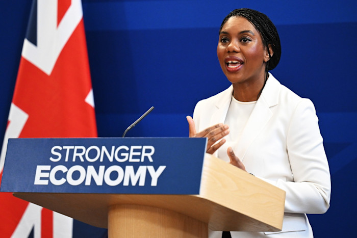 Kemi Badenoch speaks at a podium reading "Stronger Economy" with a UK flag and blue backdrop behind her.