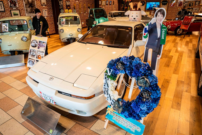 A visitor views the white Toyota Supra used by Sanae Takaichi in the 1990s, which is displayed next to a cardboard cut-out of Takaichi and a blue floral wreath.