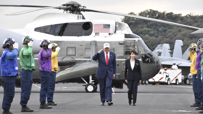 Donald Trump salutes crew members while walking alongside Sanae Takaichi on the USS George Washington, with Marine One and saluting crew members in the background.