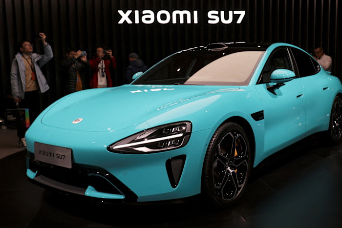 Xiaomi’s first electric vehicle, the turquoise SU7 sedan, displayed at an event as visitors take photos in the background.