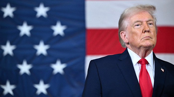 Donald Trump stands in front of a large American flag, wearing a navy suit and red tie, looking serious.