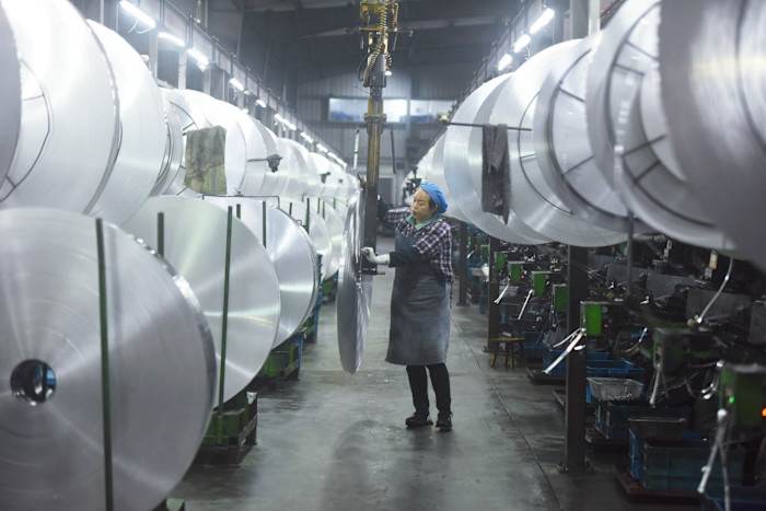 A worker operates machinery to move large rolls of aluminium material between rows of metal coils in a factory workshop.