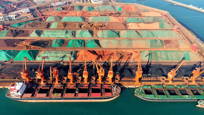 Aerial view of cargo ships docked at Qingdao Port as cranes unload iron ore onto large, colourful stockpiles at the terminal.