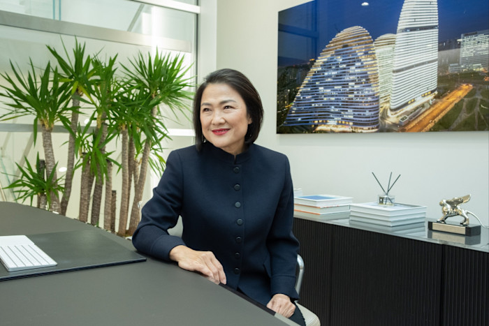Zhang Xin sits at her office desk, smiling, with a modern building photo and green plants in the background.
