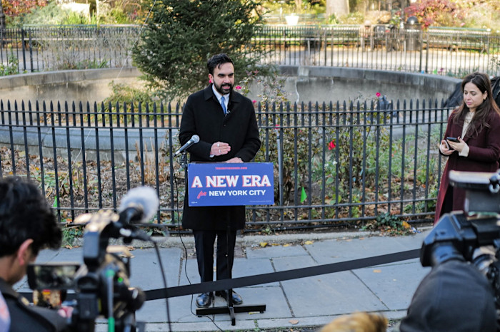 Zohran Mamdani speaks at an outdoor podium labelled ‘A New Era for New York City’ as cameras record and reporters look on.