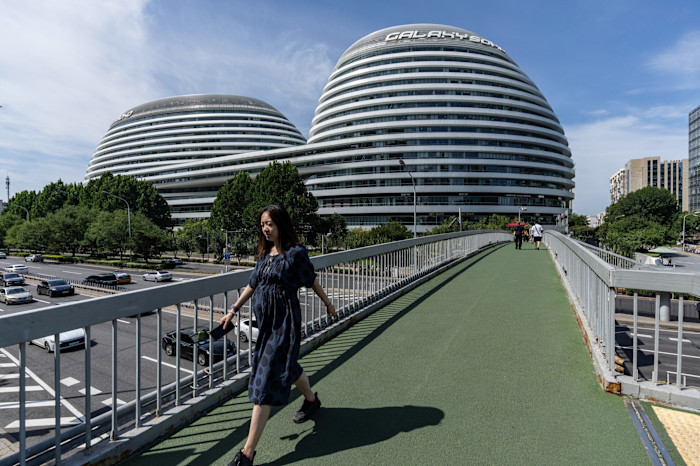 Pedestrians cross a footbridge in front of the rounded, modern Galaxy Soho complex in Beijing under a clear sky.