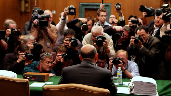 Lloyd Blankfein, with his back to the camera, faced by dozens of press photographers