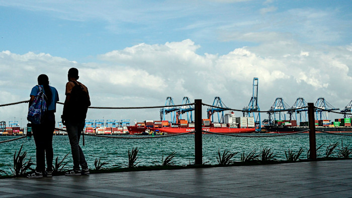 Two people stand by a waterfront railing watching cargo ships and cranes at Colon Port in Panama.