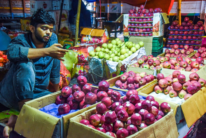 A vendor sits among crates of red and green apples at a market, looking at a mobile phone.