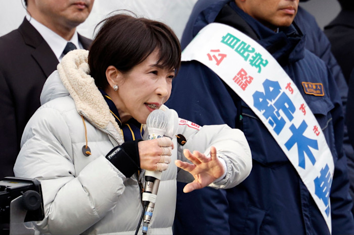 Sanae Takaichi speaks into a microphone at an election rally, gesturing with her hand, flanked by supporters.