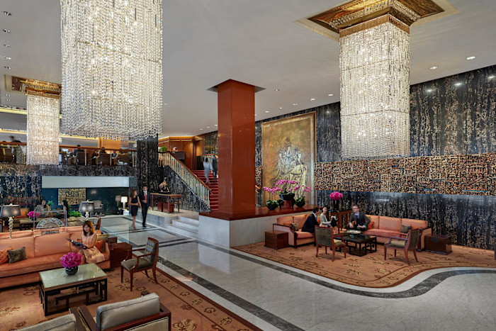The Mandarin Oriental Hong Kong’s lobby, with marble floors, large crystal chandeliers, plush seating, and guests conversing or reading. Ornate artwork and flower arrangements decorate the space.