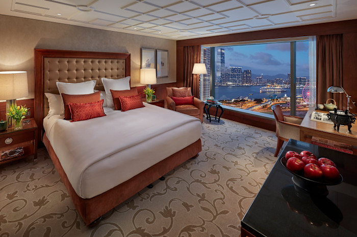 One of the hotel’s spacious Harbour View rooms, with a large bed, elegant furnishings, and a panoramic window offering a view of Hong Kong's harbor and city skyline.