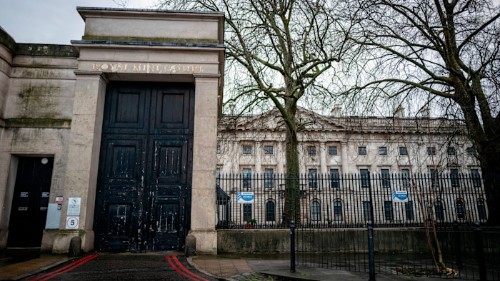 Entrance gate of Royal Mint Court with large black doors, weathered stone walls, and a historic building in the background