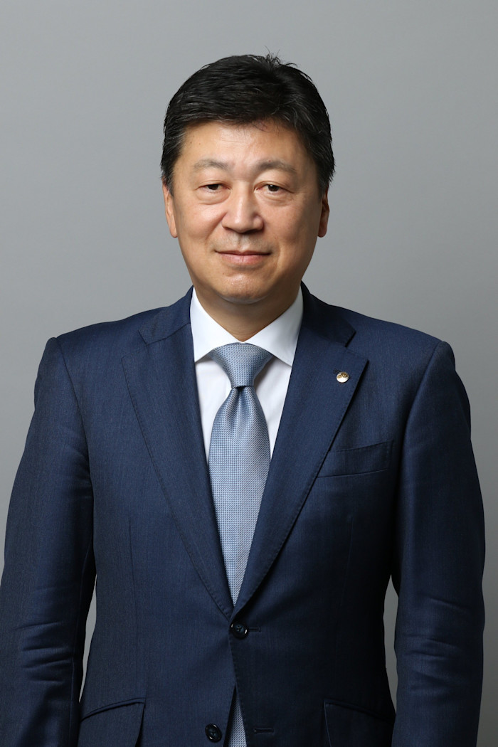 Nobuhisa Iida in a blue suit and light blue tie, standing against a plain gray background