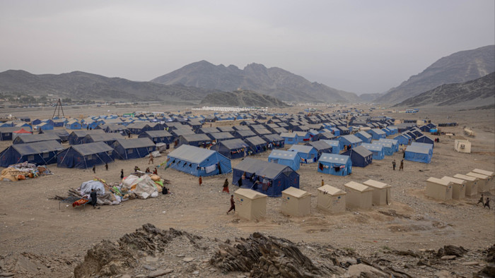 Rows of blue tents fill a barren, rocky valley at Omari Camp, with scattered people and makeshift shelters visible among them.