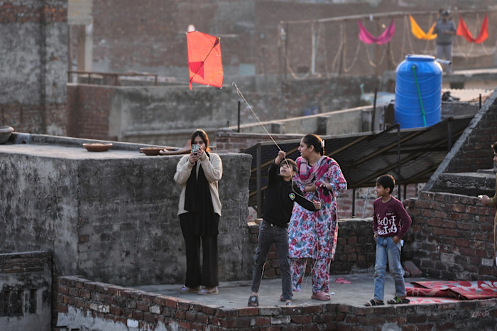 A young boy in a black top and jeans stands on a rooftop flying a red kite on a short string. At his side are a smaller boy, a girl filming him on her smartphone and a middle-aged woman in traditional floral-pattern attire.