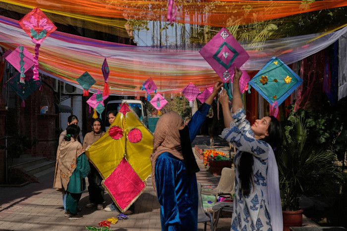 A scene in a street or courtyard with women in saris putting up overhead decorations in the shape of kites and brightly coloured drapery.