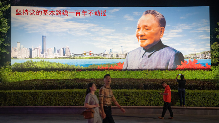 People walk past a billboard showing Deng Xiaoping with a cityscape behind him.
