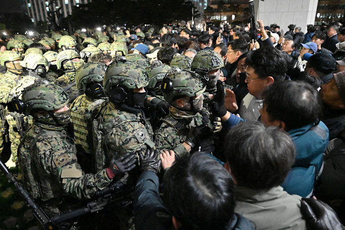 Soldiers in camouflage uniforms and helmets confront a dense crowd of civilians outside a building at night.