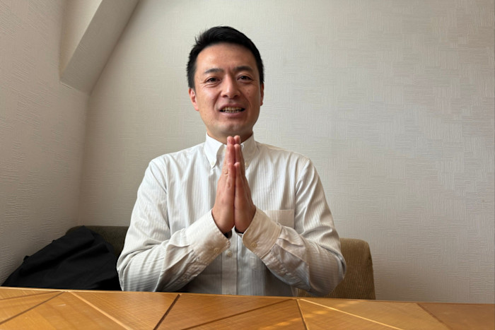 Yuichi Kondo sits at a table with his hands pressed together in front of his chest, smiling slightly.
