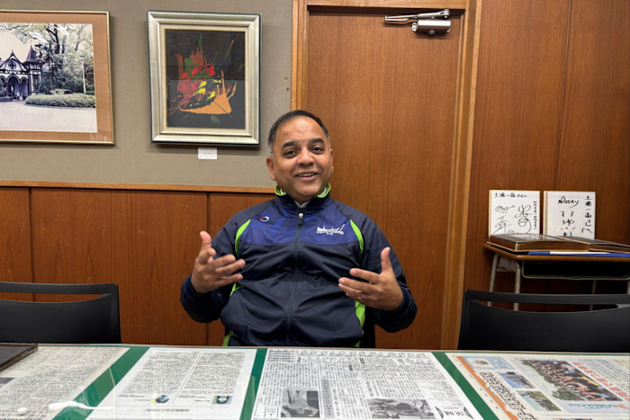 A man identified as Puranik Yogendra sits at a table covered with newspapers, gesturing with both hands and smiling.