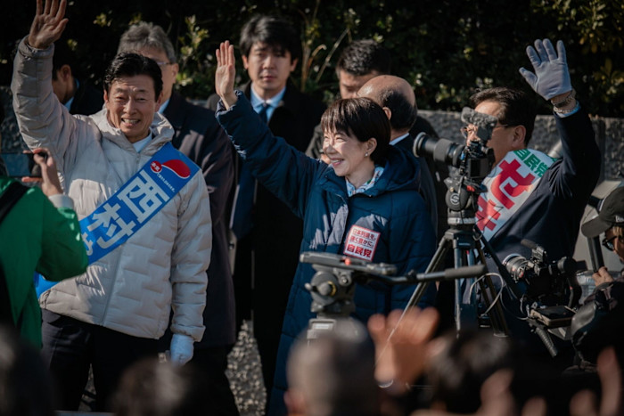 Yasutoshi Nishimura and Sanae Takaichi wave to supporters during a campaign event, surrounded by cameras and onlookers.