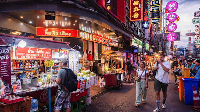 Tourists walking and browsing food stalls on a busy, brightly lit street in Bangkok’s Chinatown at night