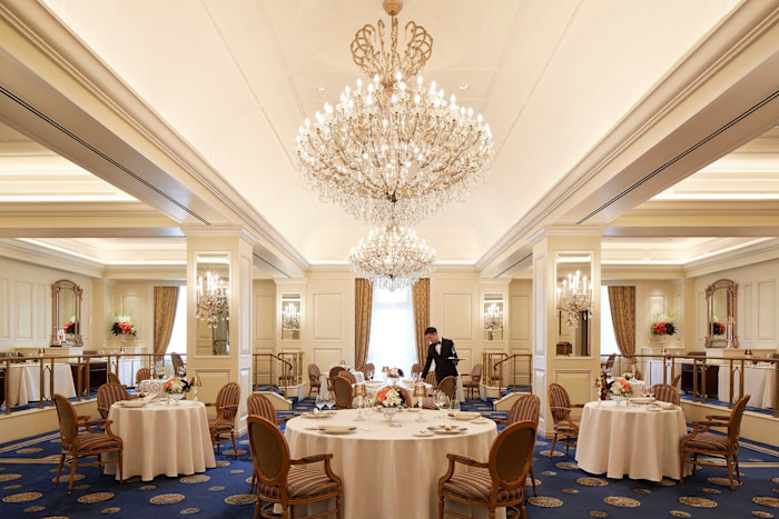 Elegant dining room with ornate chandeliers, round tables with white tablecloths and a waiter preparing a table at Gaddi’s.