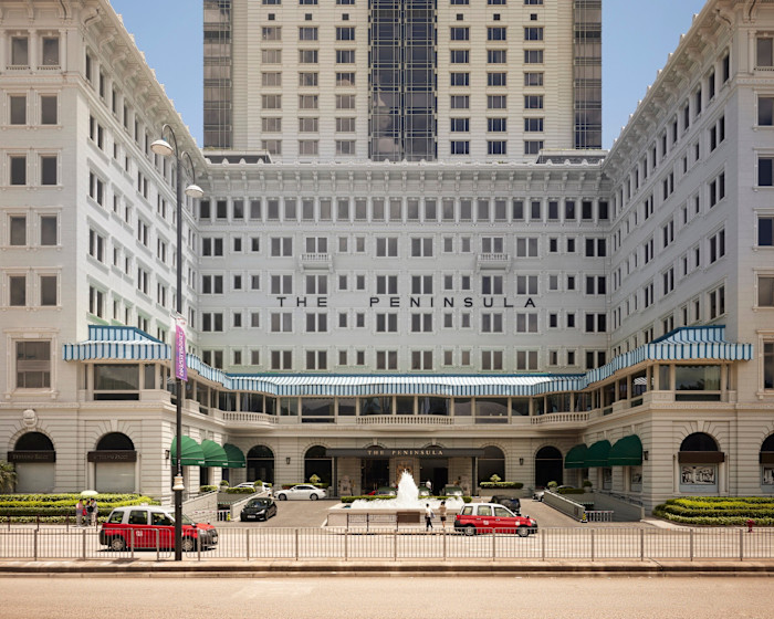 The Peninsula Hong Kong hotel exterior with white facade, blue-striped awnings, a central fountain, red taxis parked in front, and a tower behind the original six-storey building.