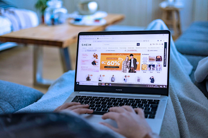 A young woman lying on a sofa under a blanket looks at the Shein online store on her laptop