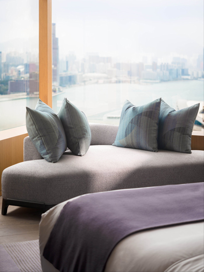 A modern bedroom in the Andre Fu Suite at Upper House, Hong Kong, featuring a grey sofa with four blue pillows and city views through large windows.