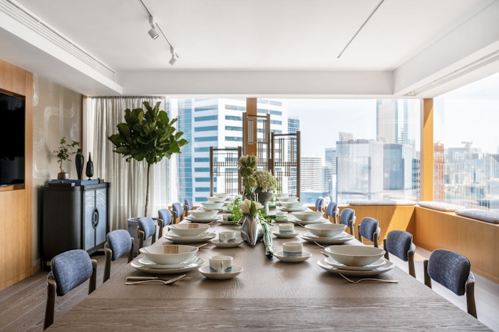 The dining room in the André Fu Suite, with a long wooden table set for 12, featuring white dishware and floral centrepieces, with city skyscrapers visible through large windows.
