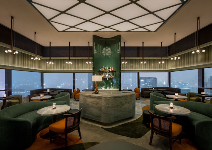 The Green Room: a stylish bar lounge with a central marble bar, green velvet seating, round tables and floor-to-ceiling windows overlooking the city.