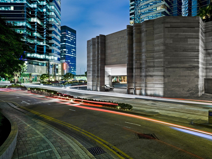 The dark-grey, windowless stone facade of Upper House Hong Kong, photographed in the evening, with long-exposure light trails running by it. Modern glass skyscrapers surround it under a deep blue twilight sky.