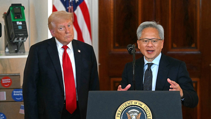 Jensen Huang speaks at a podium with the presidential seal as Donald Trump stands beside him, with flags and equipment in the background