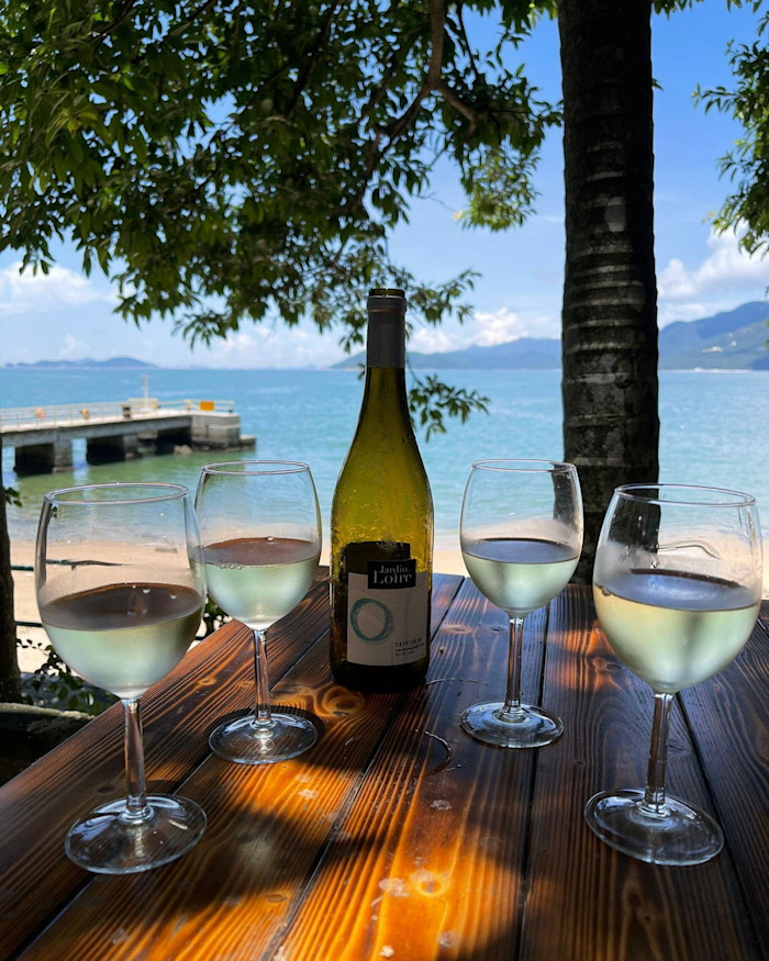 A bottle of white wine and four filled wine glasses on a wooden table, overlooking a beach and pier with blue sea and mountains at The Hong Kong Outpost on Lantau Island.