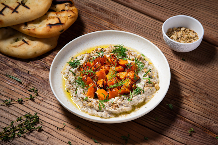 Baba ghanoush topped with roasted vegetables and herbs, served with flatbread and a bowl of dukkah on a wooden table at Pier 7.