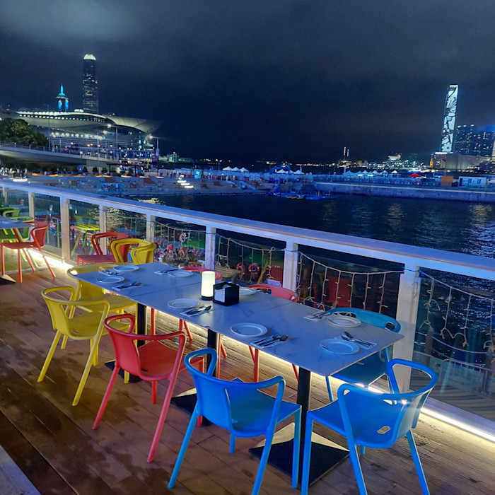 A table set for six with colourful chairs at Harbour Fun, overlooking the harbour at night, with Hong Kong’s lights and buildings in the background.