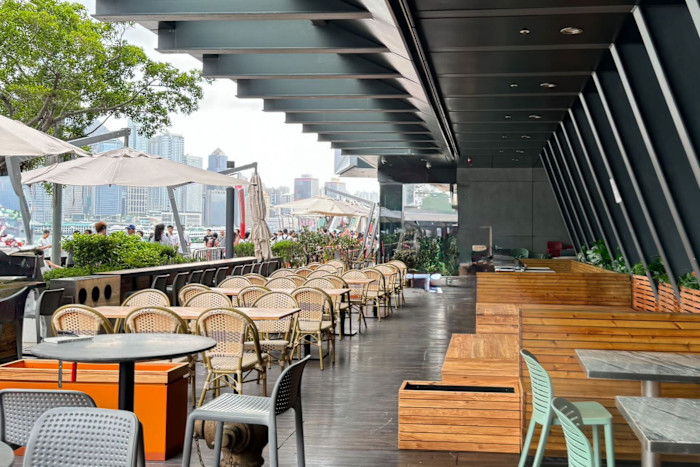 Outdoor seating area at Inki, with wooden benches, wicker chairs and tables under large umbrellas, looking across Victoria Harbour to the Central skyline