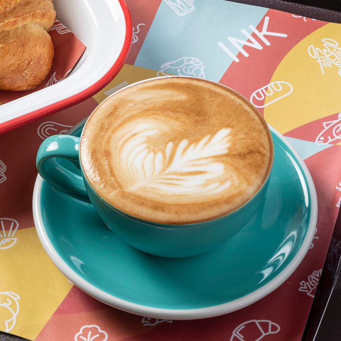 A cup of coffee with leaf-shaped art in a turquoise cup and saucer, next to a pastry on a colorful Ink-branded placemat.