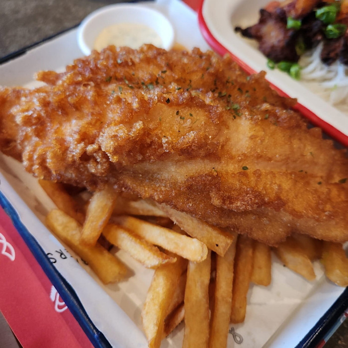 A large piece of golden fried fish served on top of thick-cut fries, with a small cup of tartar sauce on the side, at Ink.